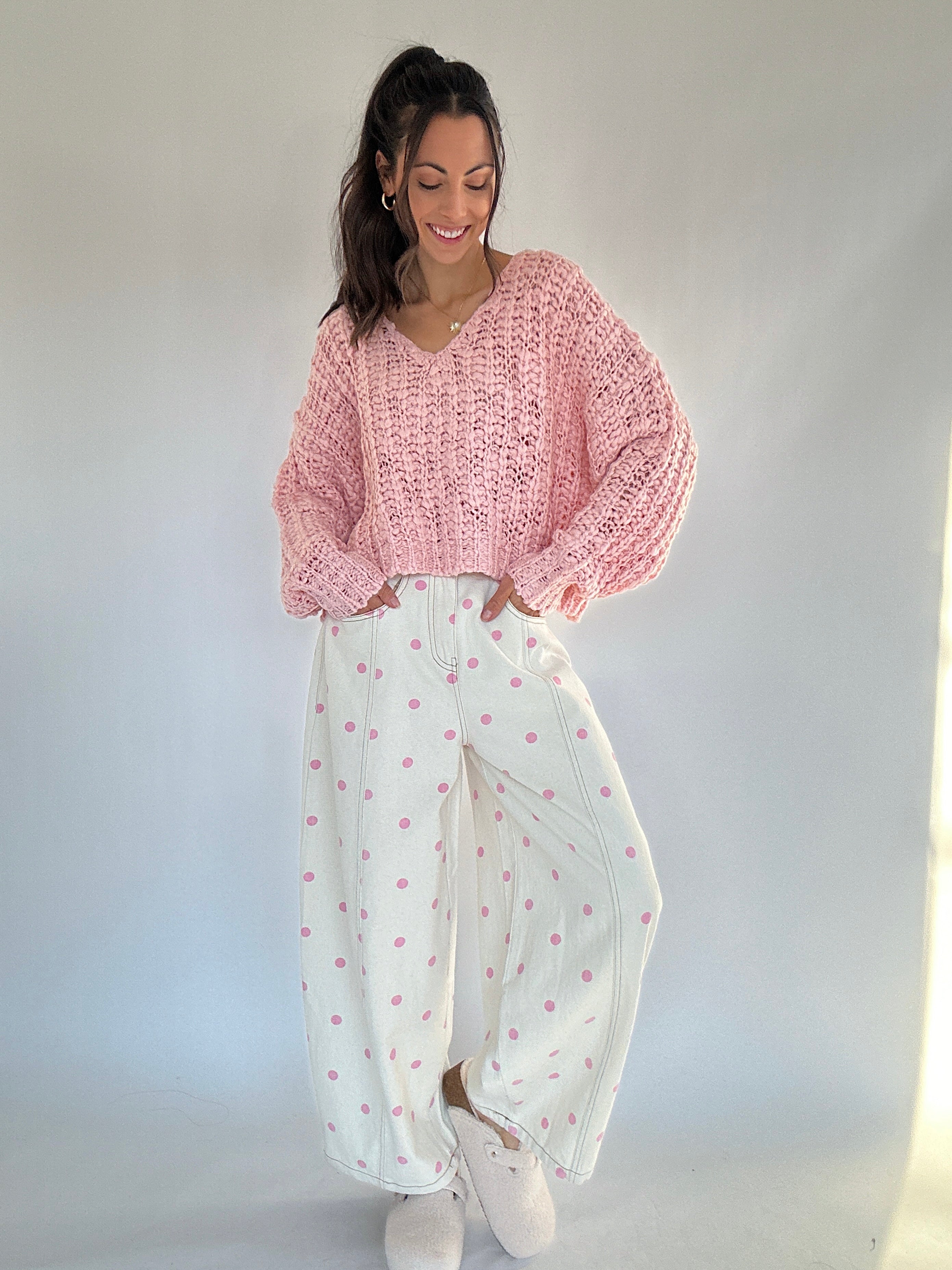 Woman wearing a pink knitted sweater and white pants with pink polka dot patterns against a plain background