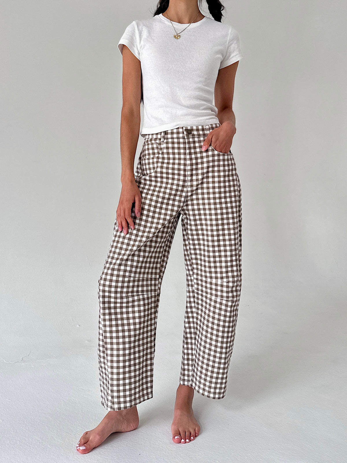 A model wearing brown and white gingham wide-leg cropped pants with a white t-shirt, standing barefoot against a light gray studio background.