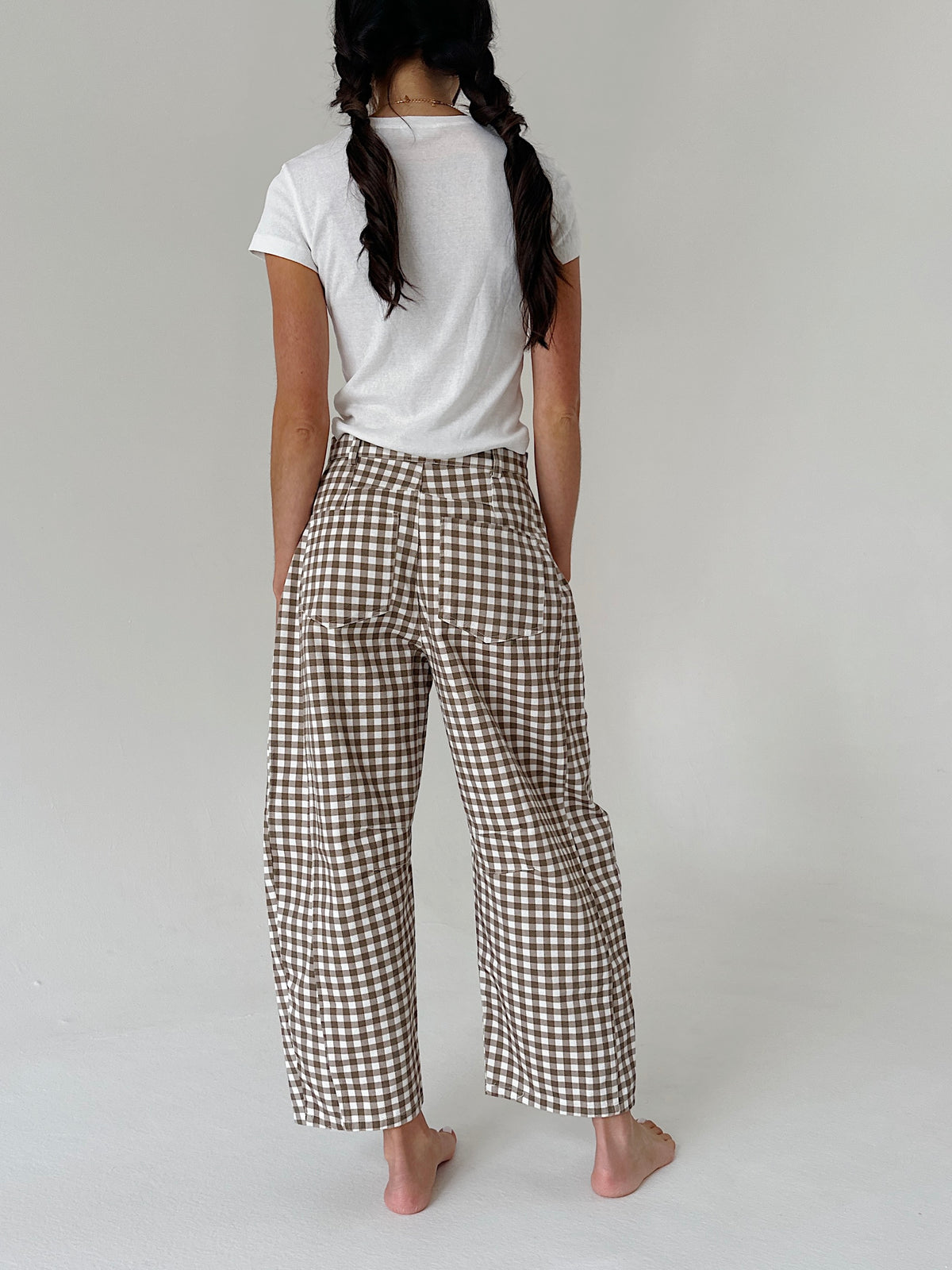 A person shown from the back wearing brown and white gingham wide-leg pants with a white t-shirt, standing barefoot against a light gray studio background.