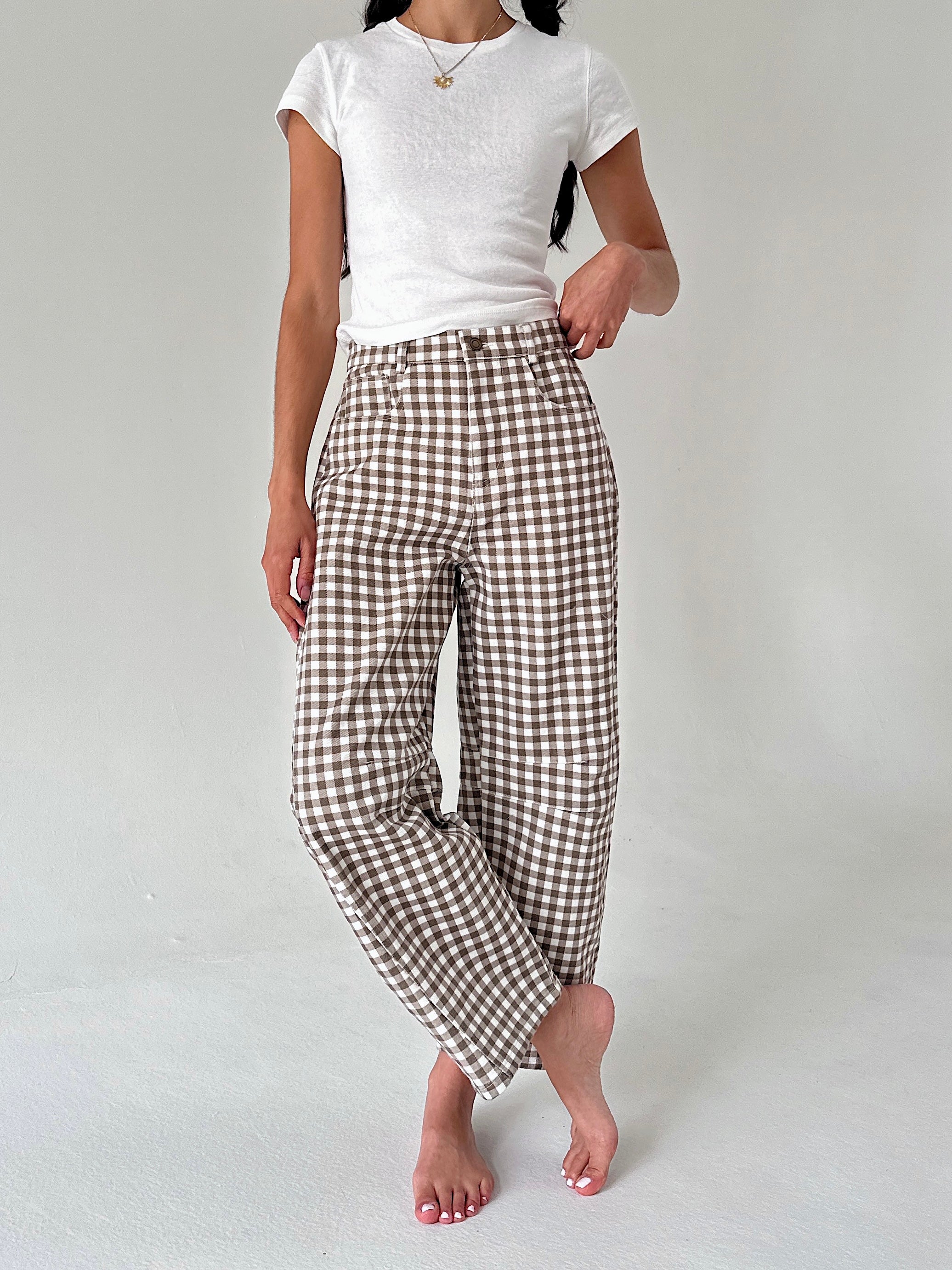 A model wearing brown and white gingham cropped trousers with a white t-shirt, standing barefoot against a light gray studio background.