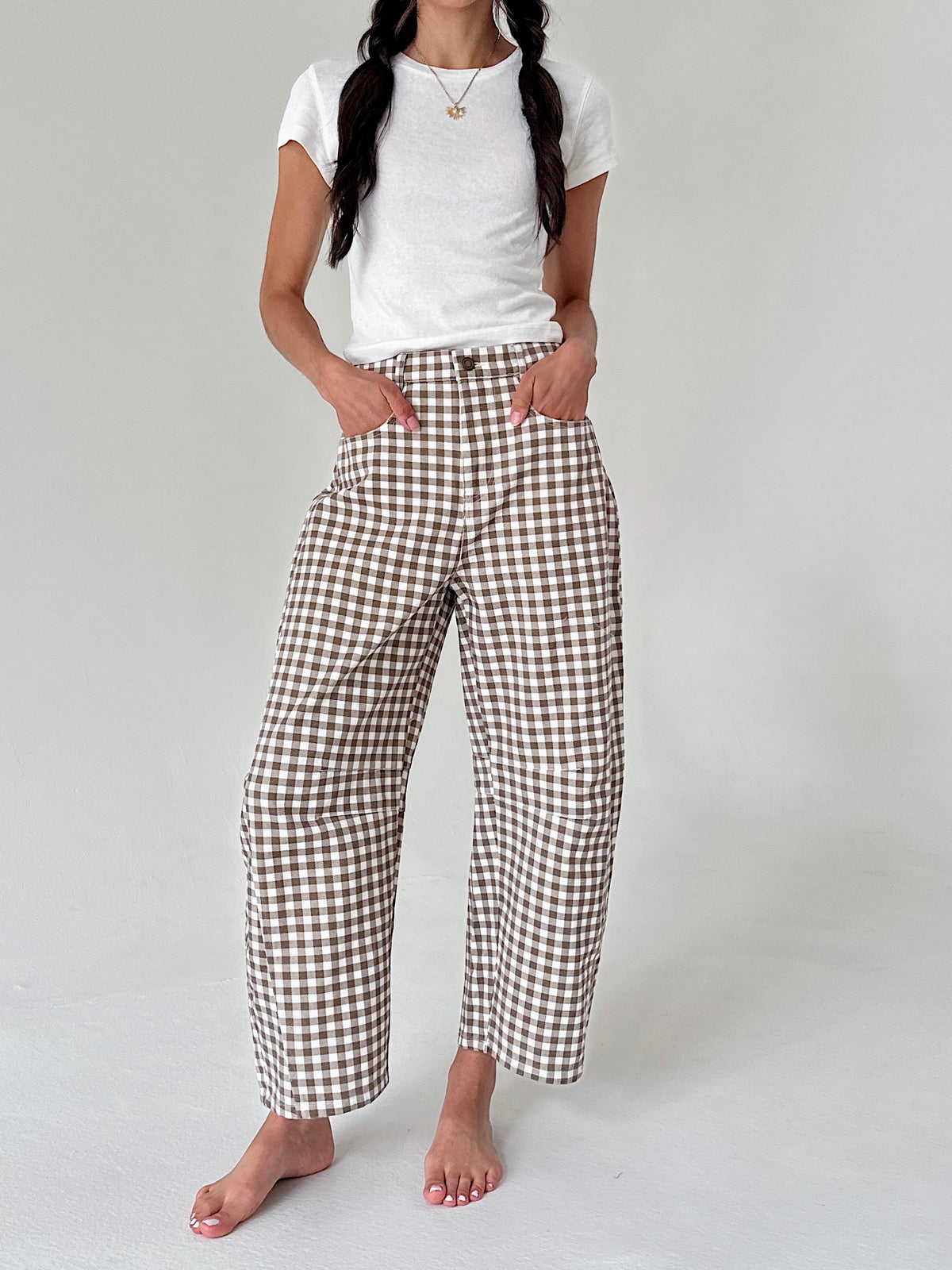 A model wearing brown and white gingham cropped wide-leg pants with a white t-shirt, standing barefoot against a light gray studio background.