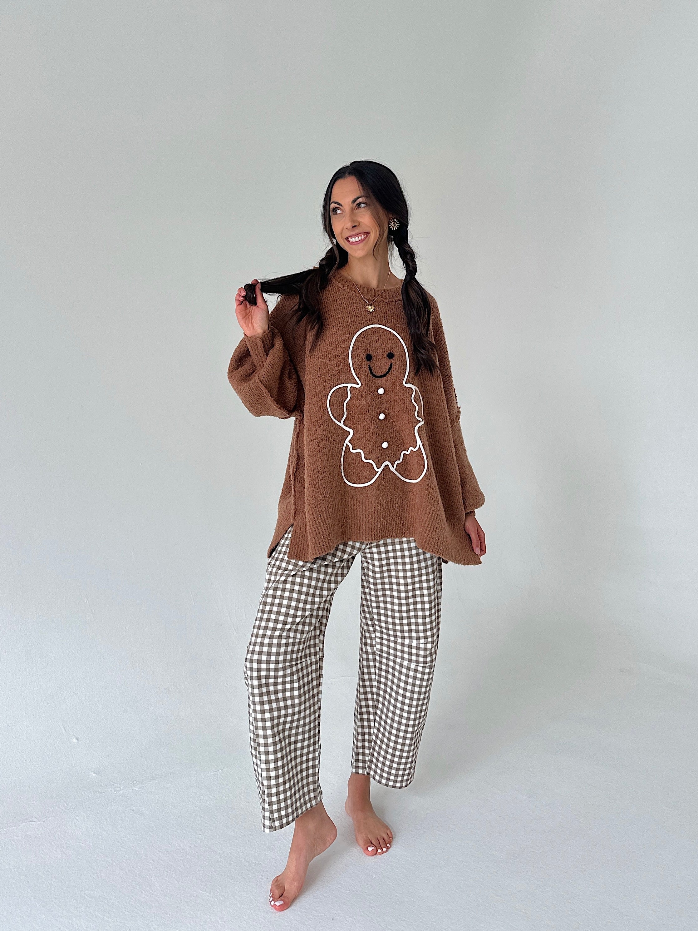 A woman modeling a cozy brown knit sweater with a white gingerbread man graphic and matching brown-and-white gingham pajama pants, standing against a light gray studio background.