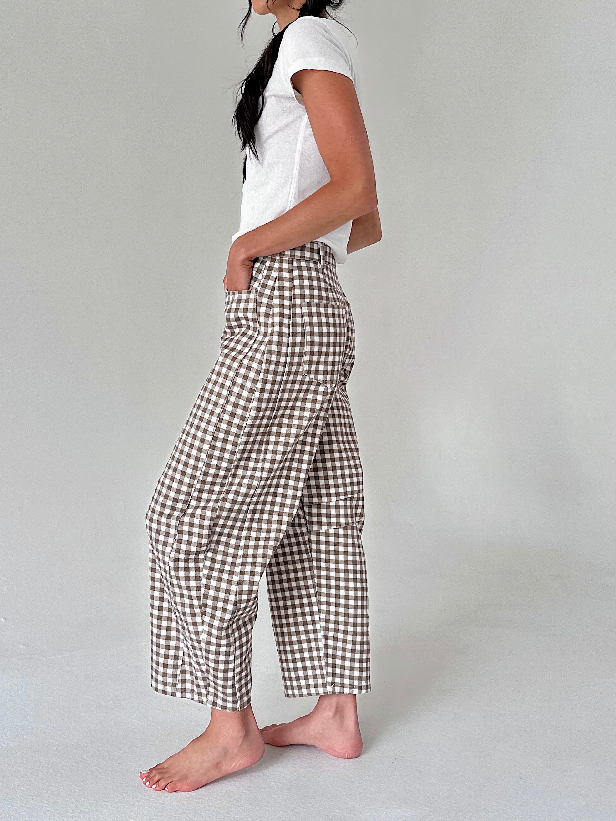 A woman wearing brown and white gingham cropped pants, a white t-shirt, and barefoot, standing against a light gray studio background.