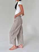 A woman wearing brown and white gingham cropped pants, a white t-shirt, and barefoot, standing against a light gray studio background.