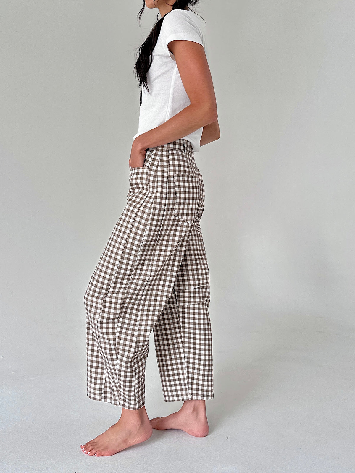 A woman wearing brown and white gingham cropped pants, a white t-shirt, and barefoot, standing against a light gray studio background.