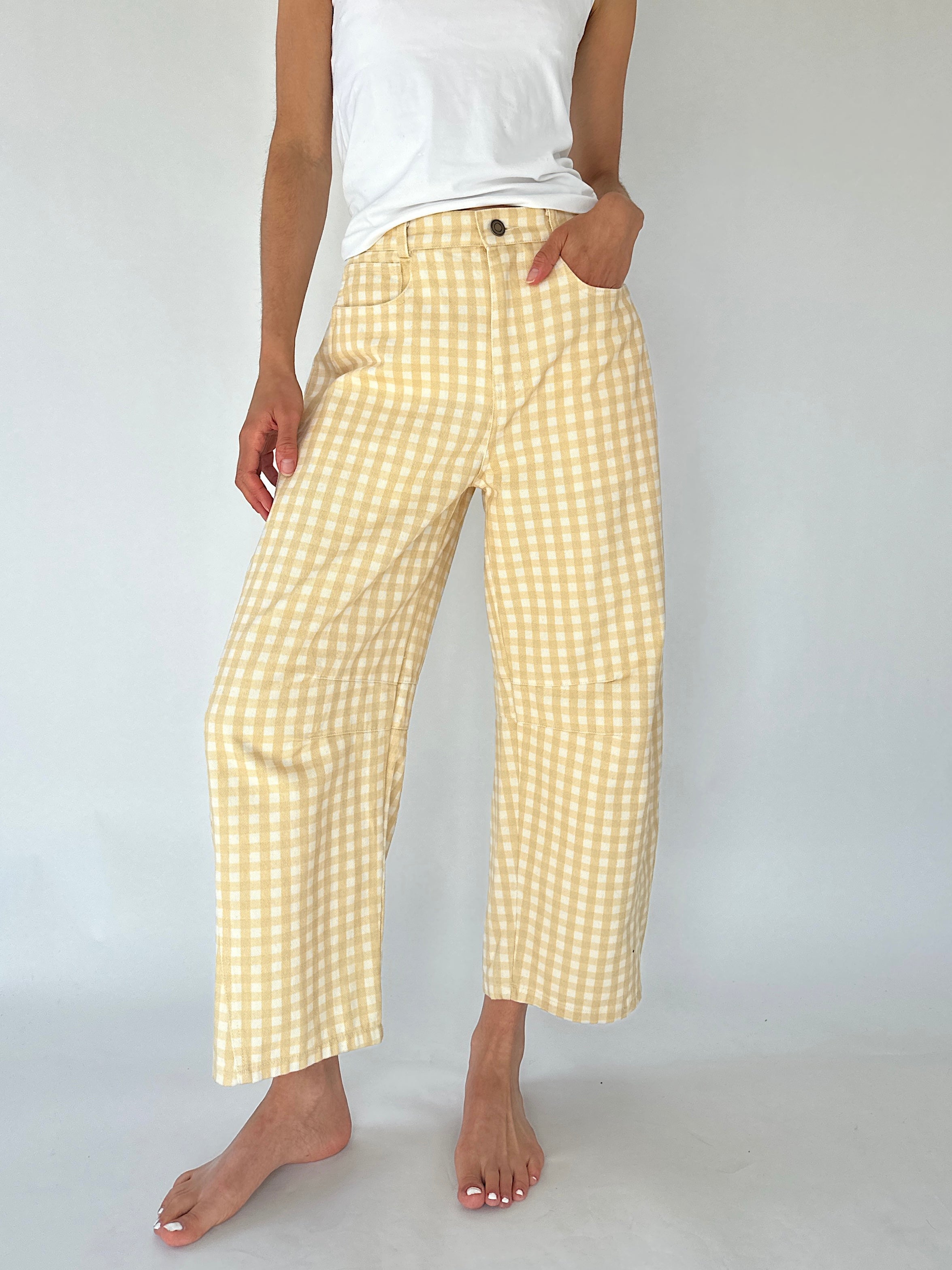 A person wearing light yellow and white gingham cropped barrel pants with a white tank top, standing barefoot against a plain light gray studio background.