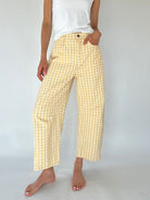 A person wearing light yellow and white gingham cropped barrel pants with a white tank top, standing barefoot against a plain light gray studio background.