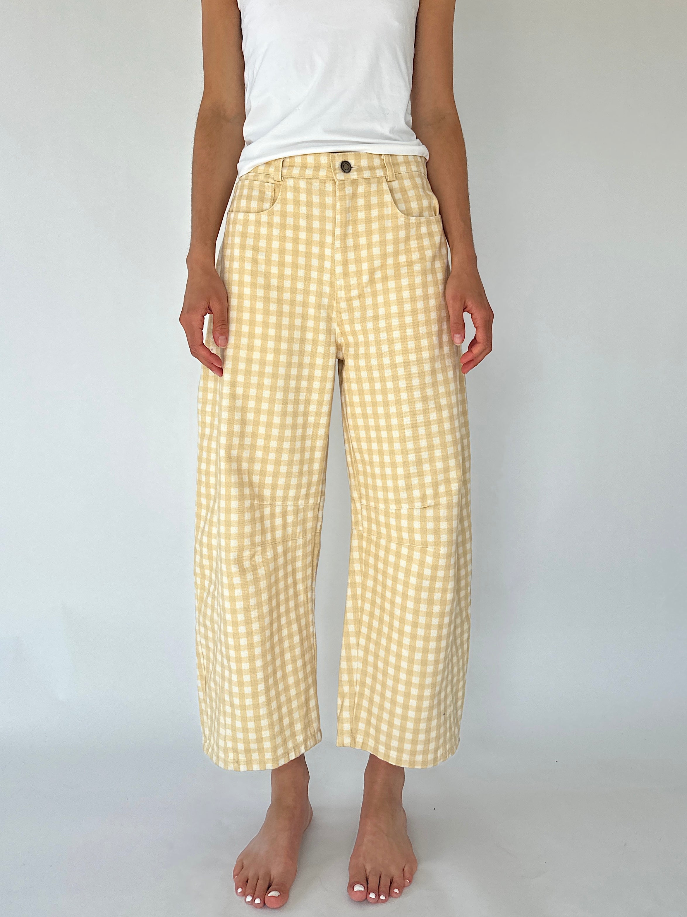 A person wearing light yellow and white gingham cropped pants with a white sleeveless top, standing barefoot against a plain light gray studio background.