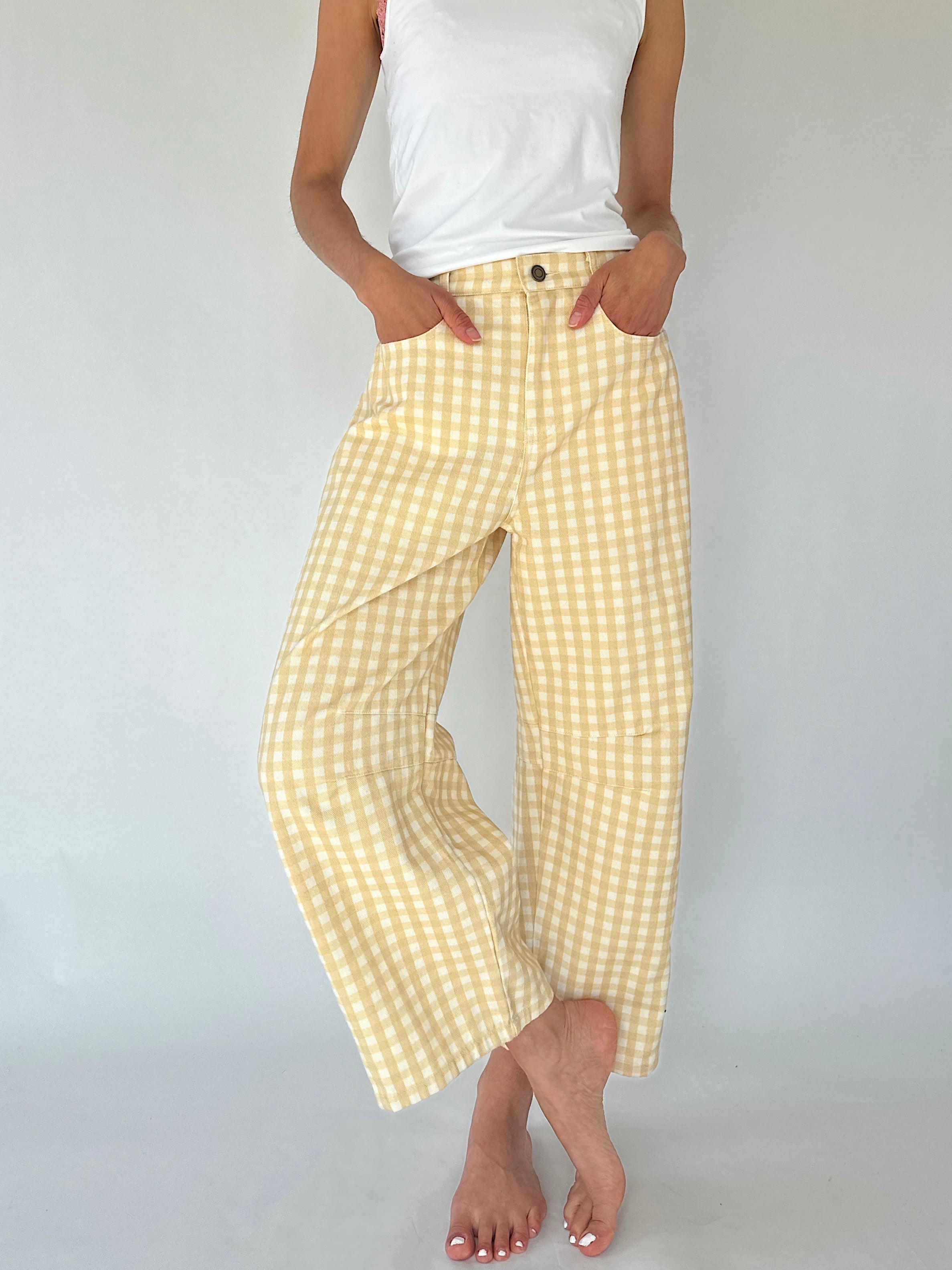 A person wearing loose, wide-leg cropped pants in a pale yellow and white gingham check pattern, paired with a white tank top, standing against a light gray studio background.
