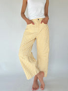 A person wearing loose, wide-leg cropped pants in a pale yellow and white gingham check pattern, paired with a white tank top, standing against a light gray studio background.