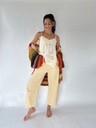 A woman stands barefoot against a plain light gray studio background, wearing a cream lace camisole, cream gingham cropped pants, and a chunky multicolor knit cardigan draped over her arms.