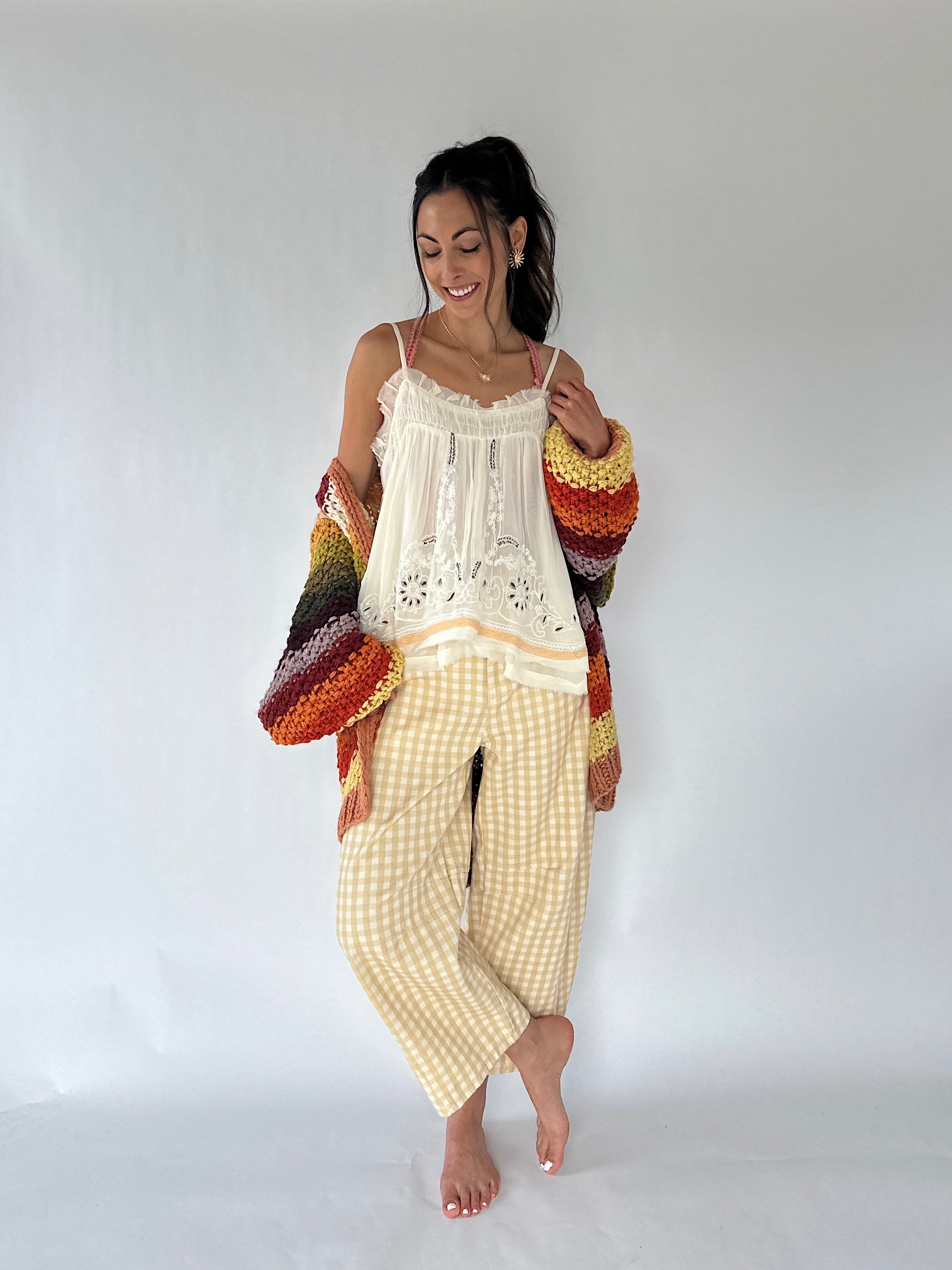A woman modeling a bohemian-style two-piece pajama set consisting of a cream lace camisole top and yellow-and-white gingham pants, styled with a multicolor striped knit cardigan, against a plain white studio background.