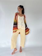 A woman modeling a cozy bohemian-style outfit consisting of a multicolor striped crochet cardigan and a cream crochet top paired with yellow-and-white gingham cropped pants, against a plain white studio background.