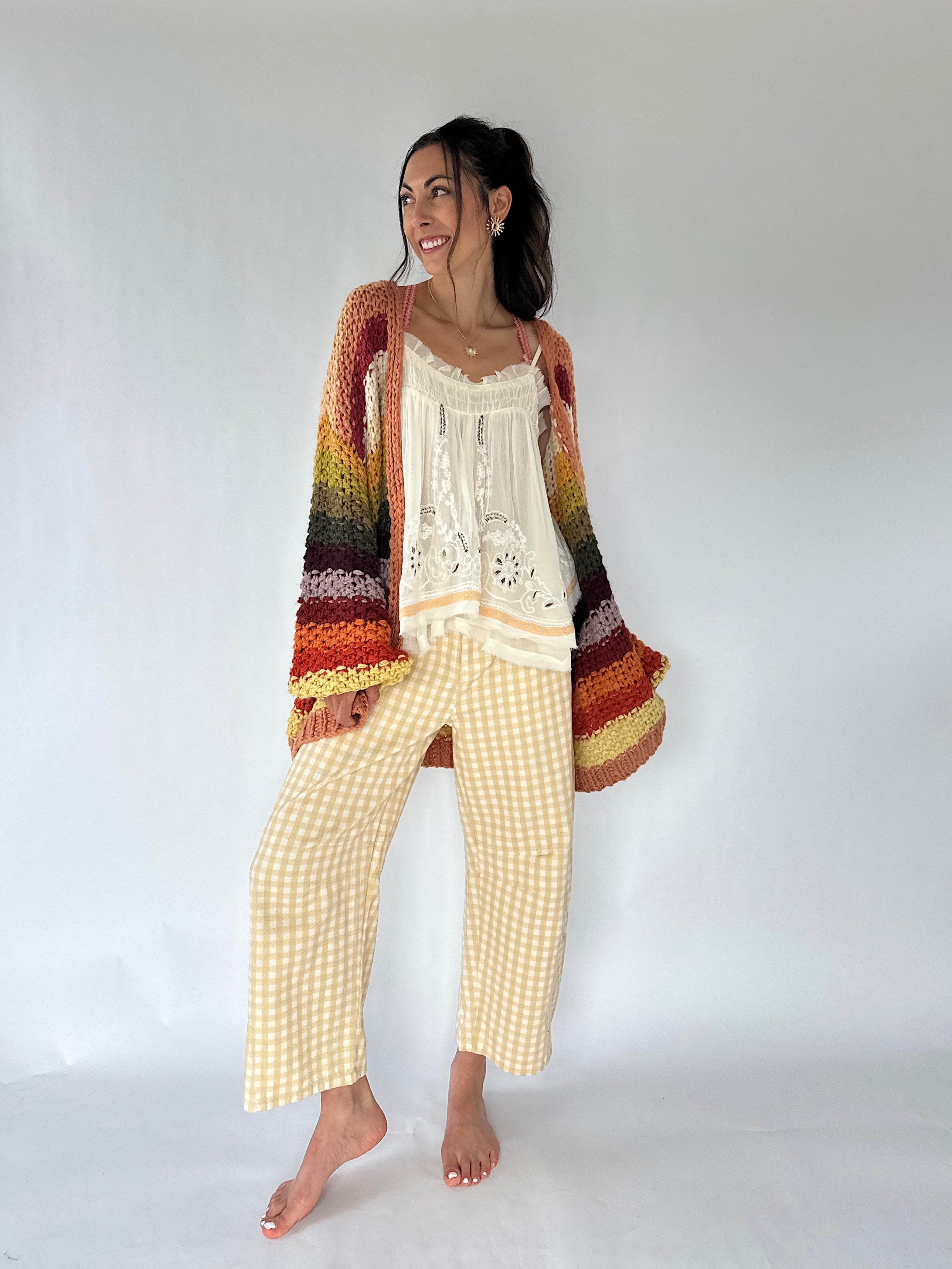 A woman modeling a cozy bohemian-style outfit consisting of a multicolor striped crochet cardigan and a cream crochet top paired with yellow-and-white gingham cropped pants, against a plain white studio background.
