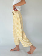 A person wearing loose, barrel cropped pants in a pale yellow and white gingham check pattern, paired with a white top, shown from the waist down against a light gray studio background.