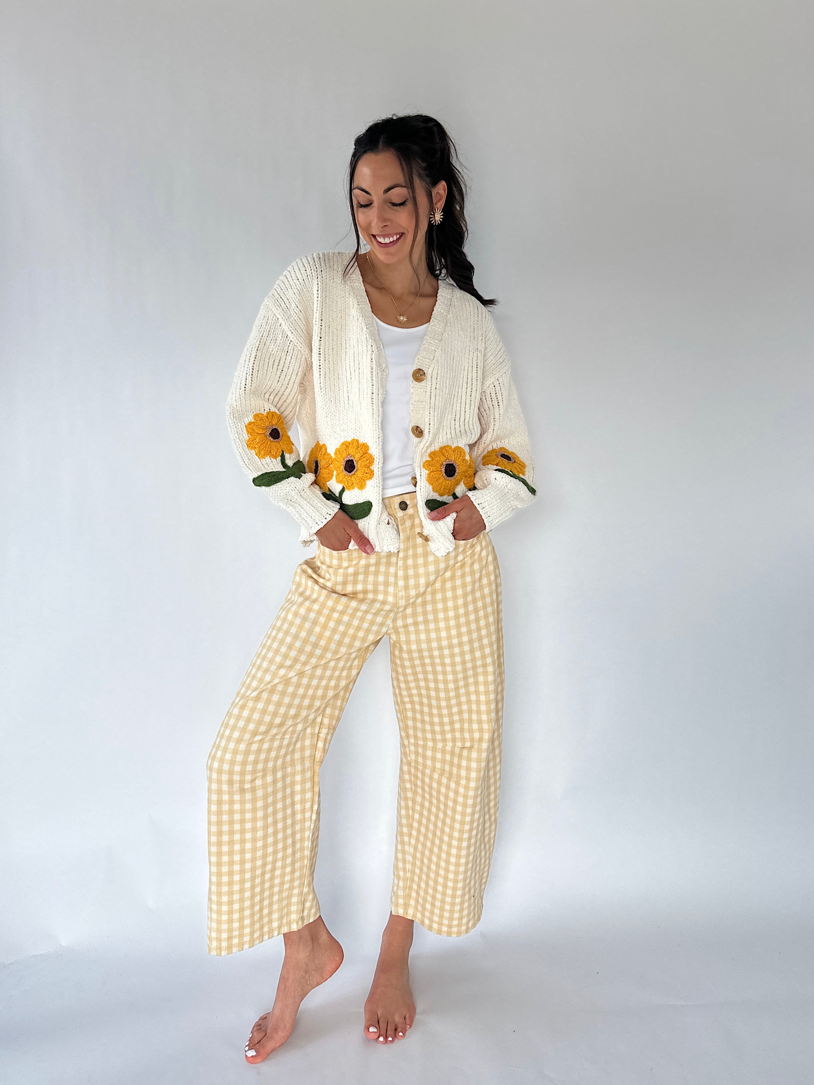 A woman stands barefoot against a plain light gray studio background, wearing a cream knit cardigan with embroidered sunflowers and yellow-and-white gingham cropped pants.