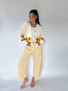 A woman stands barefoot against a plain light gray studio background, wearing a cream knit cardigan with embroidered sunflowers and yellow-and-white gingham cropped pants.