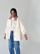 A woman modeling a cream-colored utility jacket with red-and-white gingham trim on the collar and cuffs, worn over a white t-shirt and blue jeans against a plain light gray background.