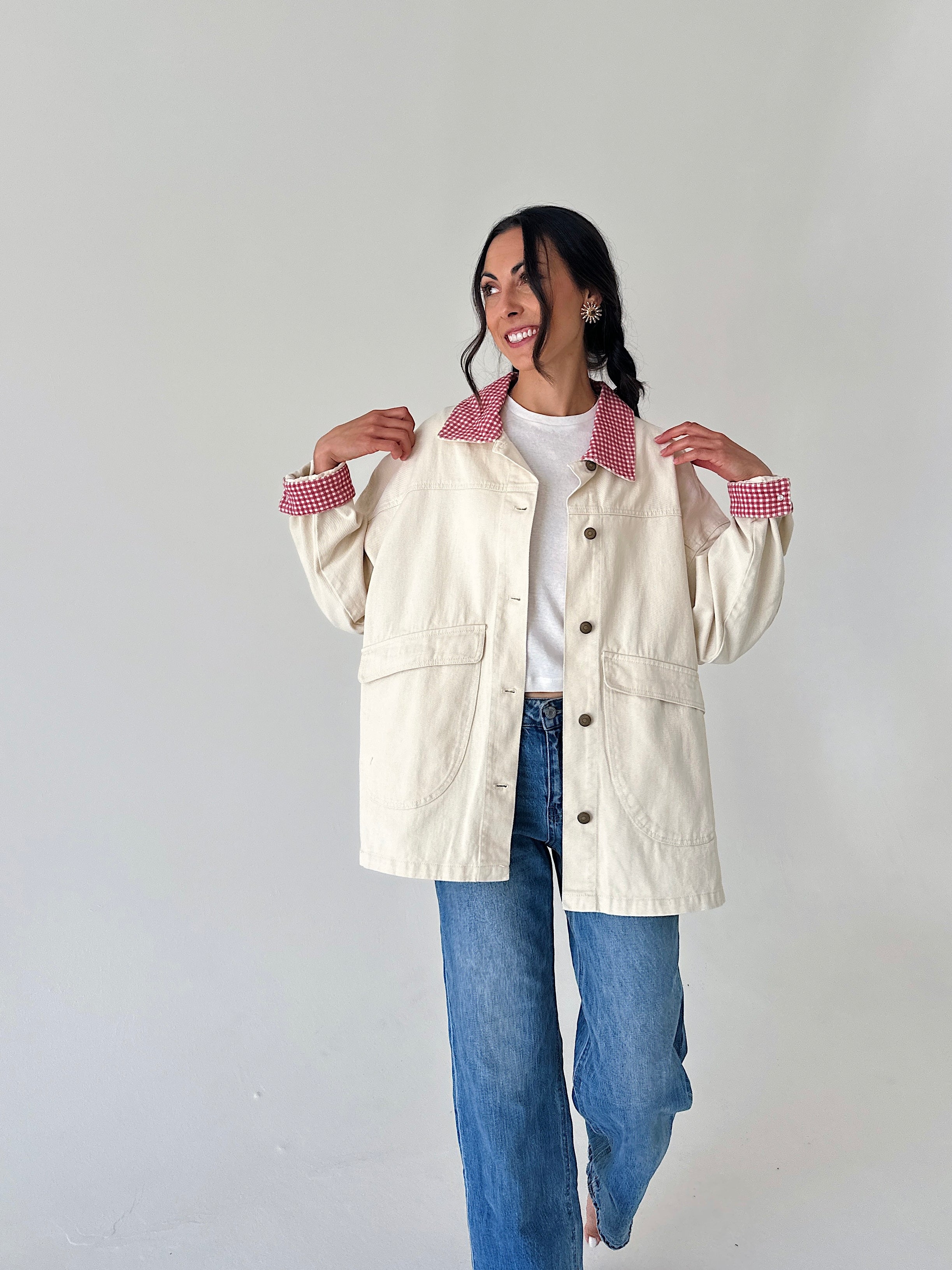 A woman modeling a cream-colored utility jacket with red-and-white gingham patterned collars and cuffs, worn over a white t-shirt and blue jeans, against a plain light gray background.