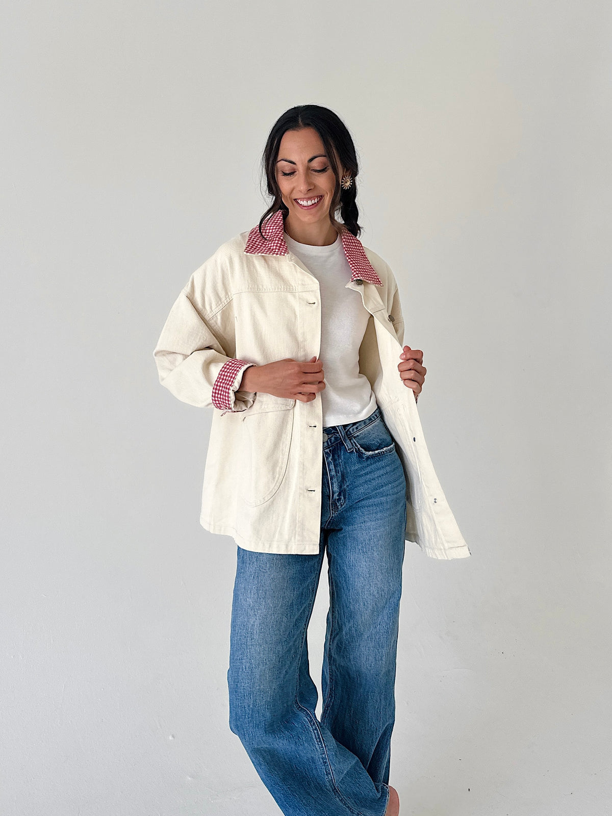 A woman modeling a cream-colored utility jacket with a pink and white gingham patterned collar and cuffs, worn over a white t-shirt and blue jeans, against a plain light gray background.