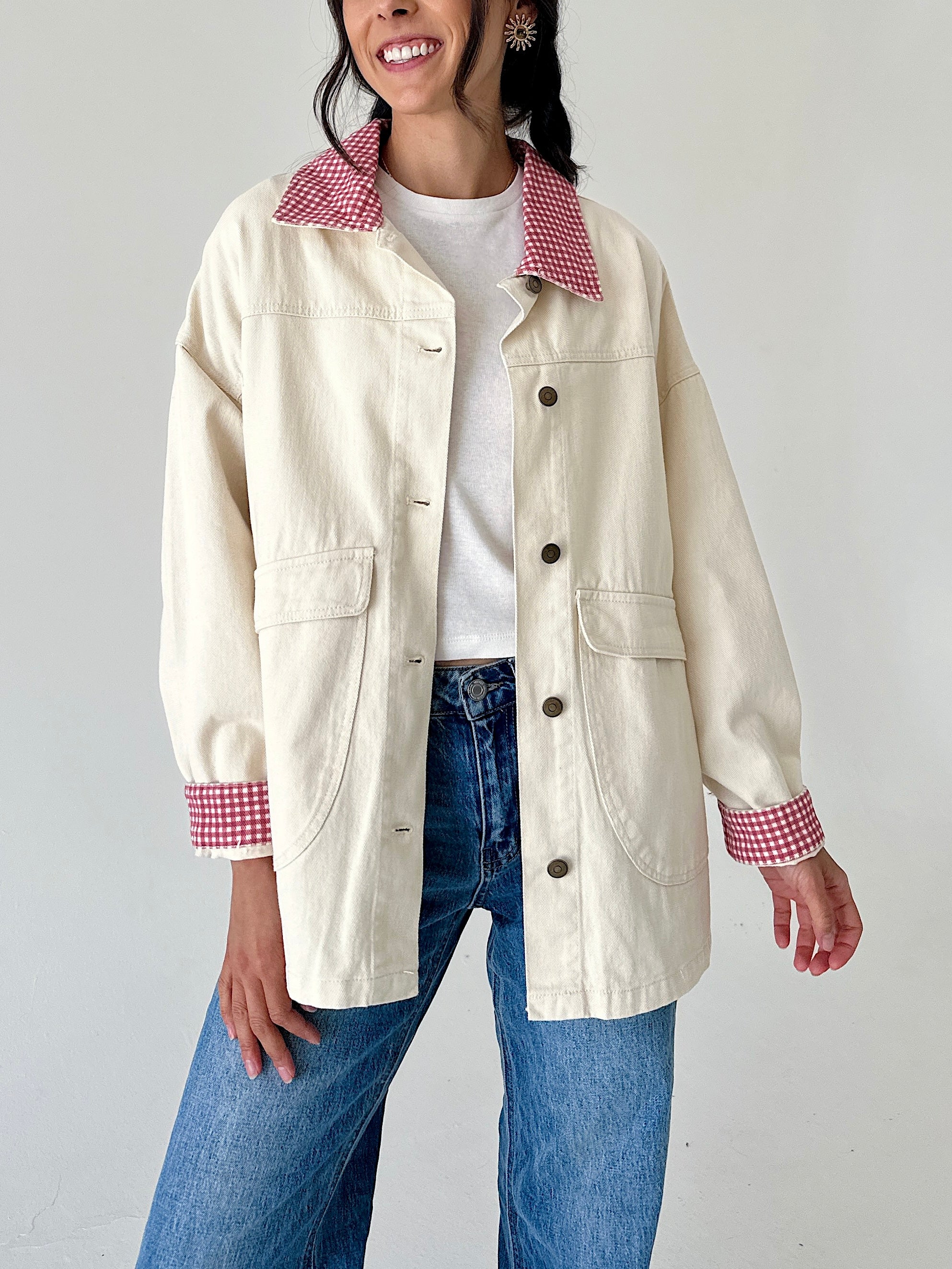 A woman wearing a cream-colored utility jacket with red and white gingham patterned collar and cuffs, paired with a white t-shirt and blue jeans.
