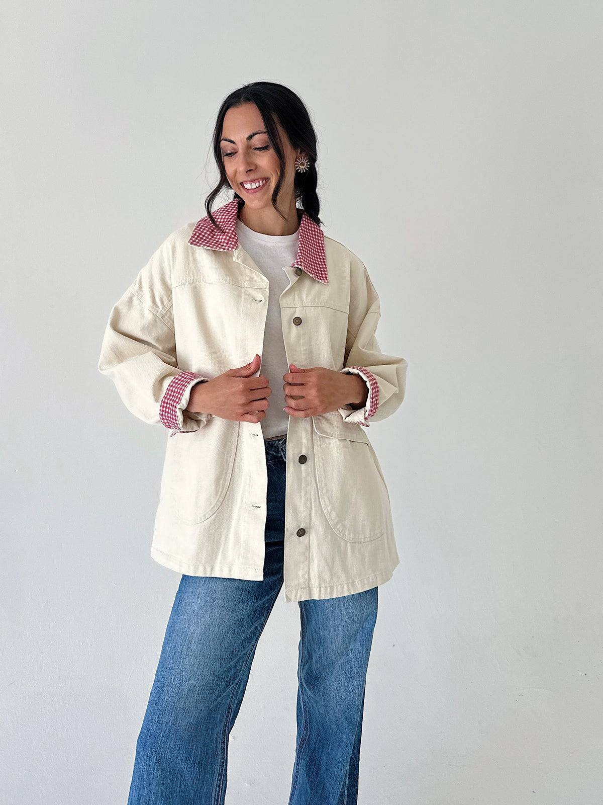 A woman modeling a cream-colored utility jacket with a red-and-white gingham patterned collar and matching gingham turned-back cuffs, paired with blue jeans against a plain light gray background.