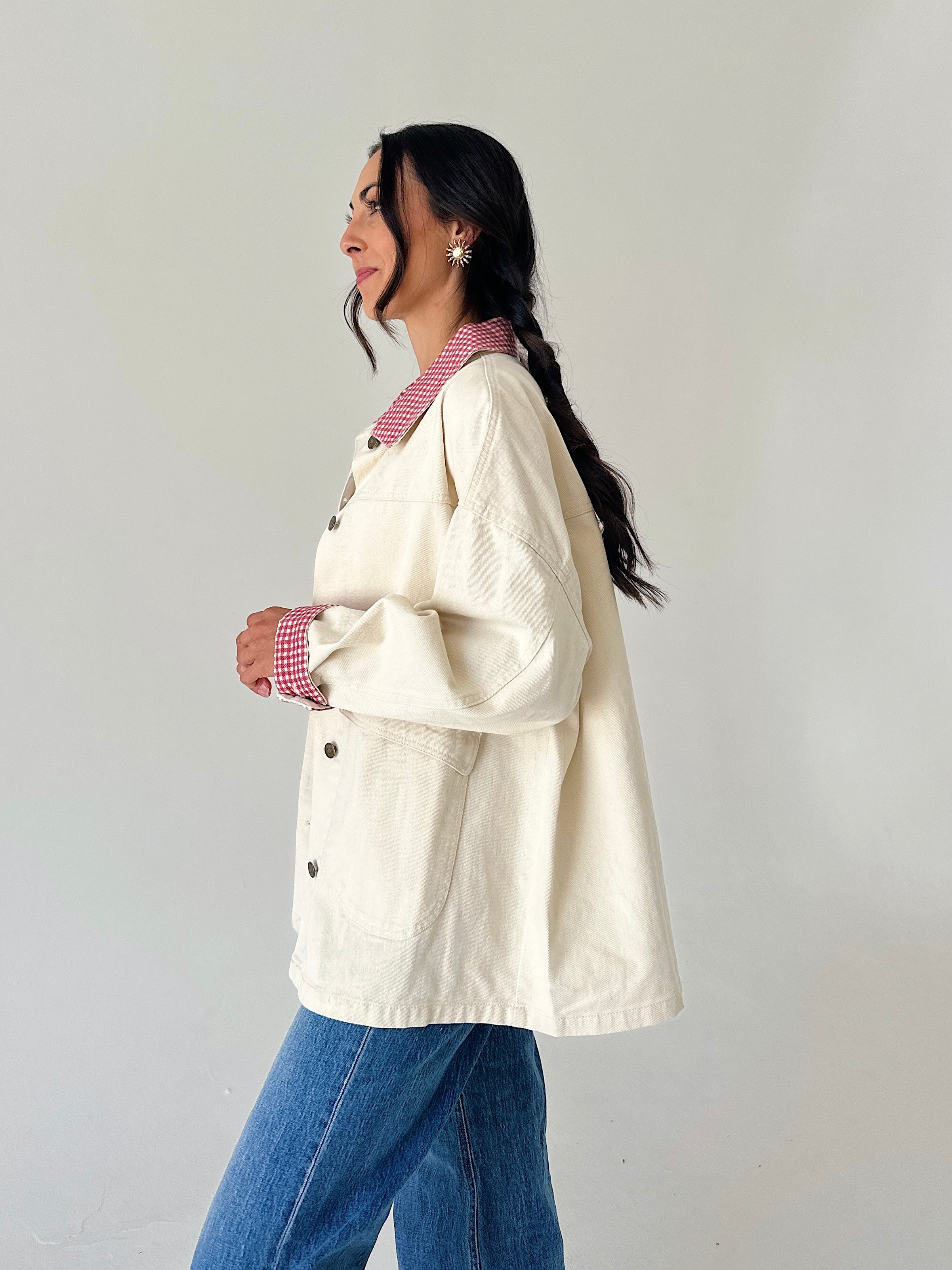 A woman wearing an oversized cream-colored utility jacket with red-and-white gingham trim on the collar and cuffs, layered over a red-and-white gingham shirt and paired with blue jeans, shown in profile against a plain light gray wall.