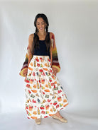 A woman modeling a long white maxi skirt with a colorful food-themed print, styled with a black crop top, a multicolor knit cardigan, and beige woven flat shoes against a plain white studio background.
