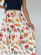 A high-waisted, tiered white skirt with a colorful food and drink print, featuring illustrations of bread, wine bottles, grapes, cherries, and baskets. The skirt is shown on a model wearing a navy blue crop top against a plain light gray background.