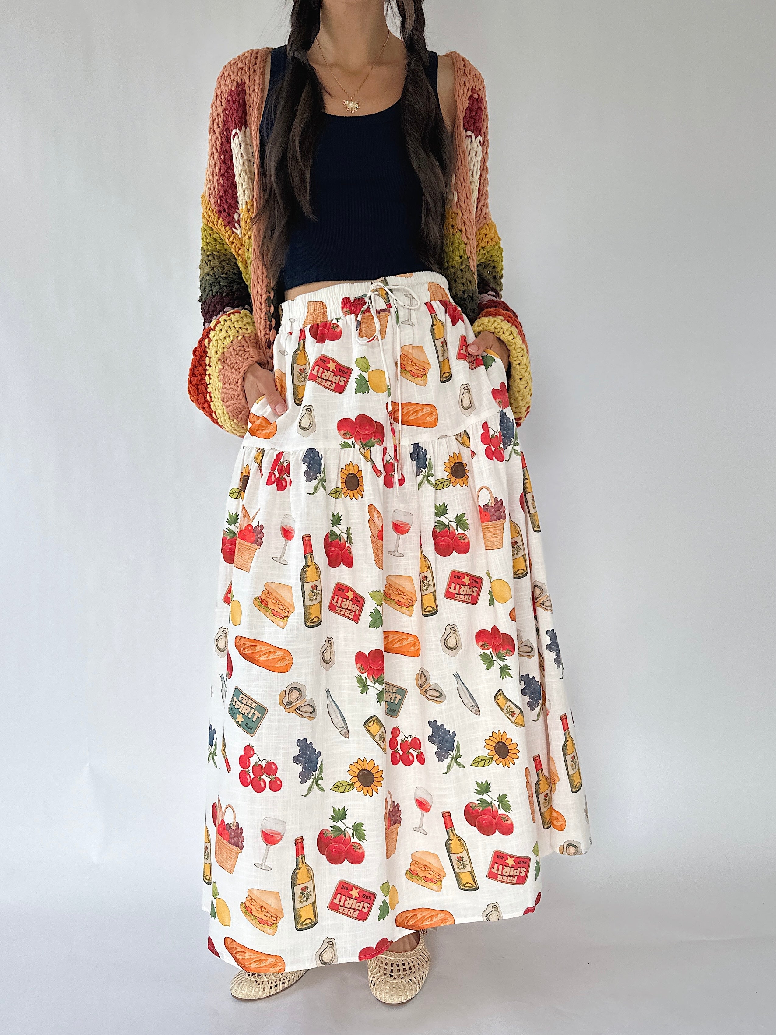 A woman wearing a long white maxi skirt with a colorful food and drink print, paired with a black top and a multicolor knit cardigan, standing against a plain light gray background.