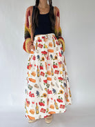 A woman wearing a long white maxi skirt with a colorful food and drink print, paired with a black top and a multicolor knit cardigan, standing against a plain light gray background.