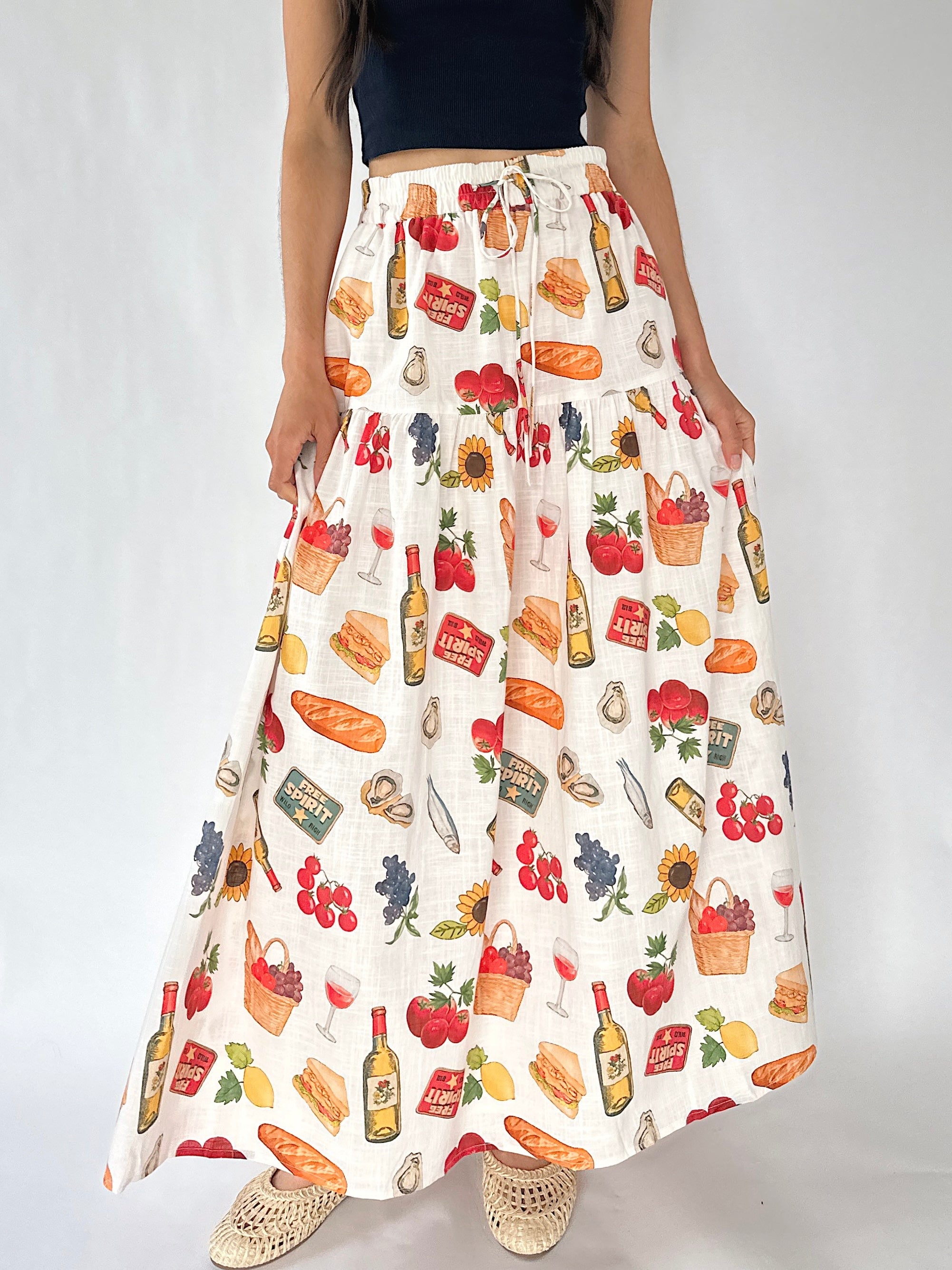 A high-waisted, wide-leg maxi skirt with a white base and a colorful print of food and drink motifs including bread, wine bottles, grapes, cherries, and baskets, modeled with a black crop top and beige woven shoes.