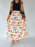 A woman models a long white skirt with a colorful wine-themed print, styled with a rainbow-striped knit cardigan, a black top, and beige woven shoes against a plain white studio background.