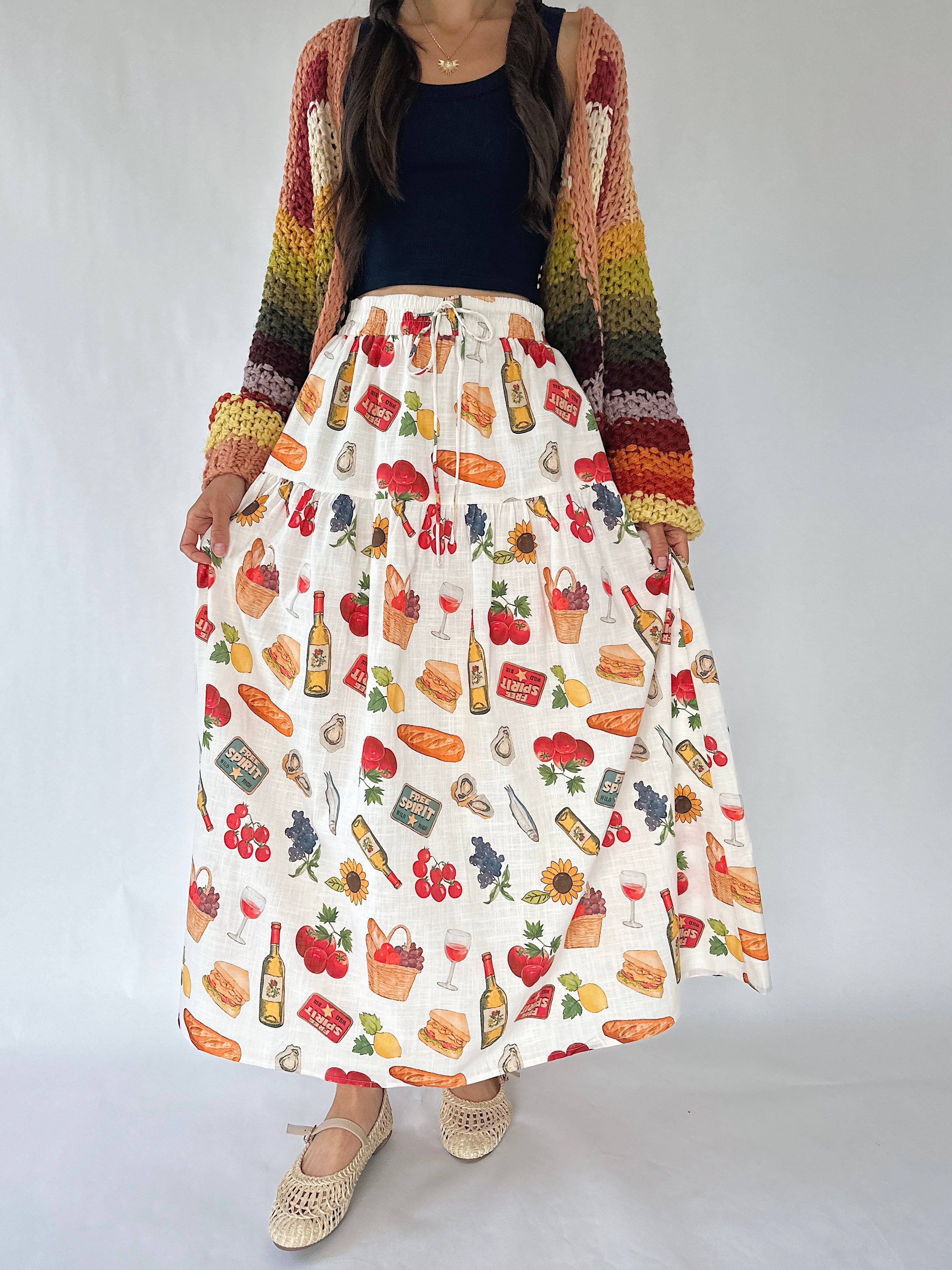 A woman models a long white skirt with a colorful wine-themed print, styled with a rainbow-striped knit cardigan, a black top, and beige woven shoes against a plain white studio background.