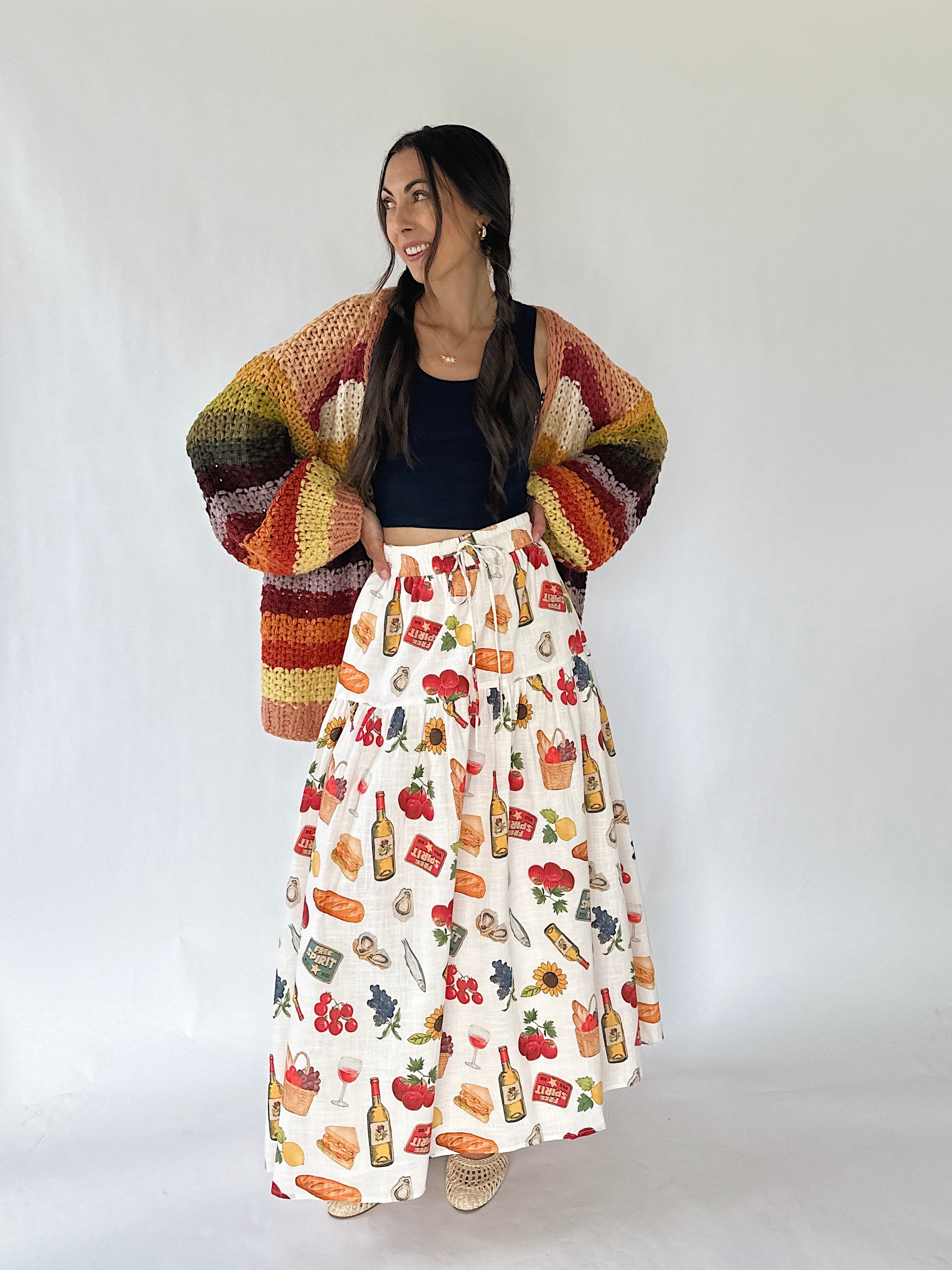 A woman modeling a colorful striped crochet cardigan over a black crop top and a long white skirt with a colorful food and drink print, standing against a plain white studio background.