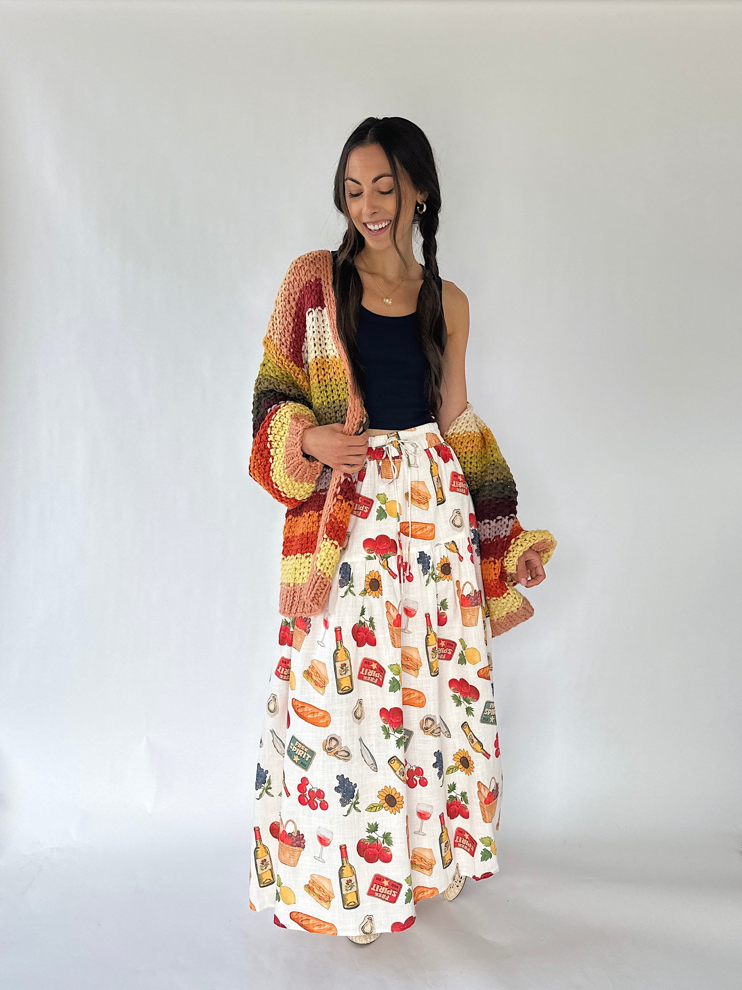 A woman modeling a long white skirt with a colorful food and drink print, paired with a dark navy top and a chunky multicolor crochet cardigan, standing against a plain white studio background.