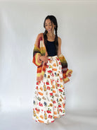 A woman modeling a long white skirt with a colorful food and drink print, paired with a dark navy top and a chunky multicolor crochet cardigan, standing against a plain white studio background.