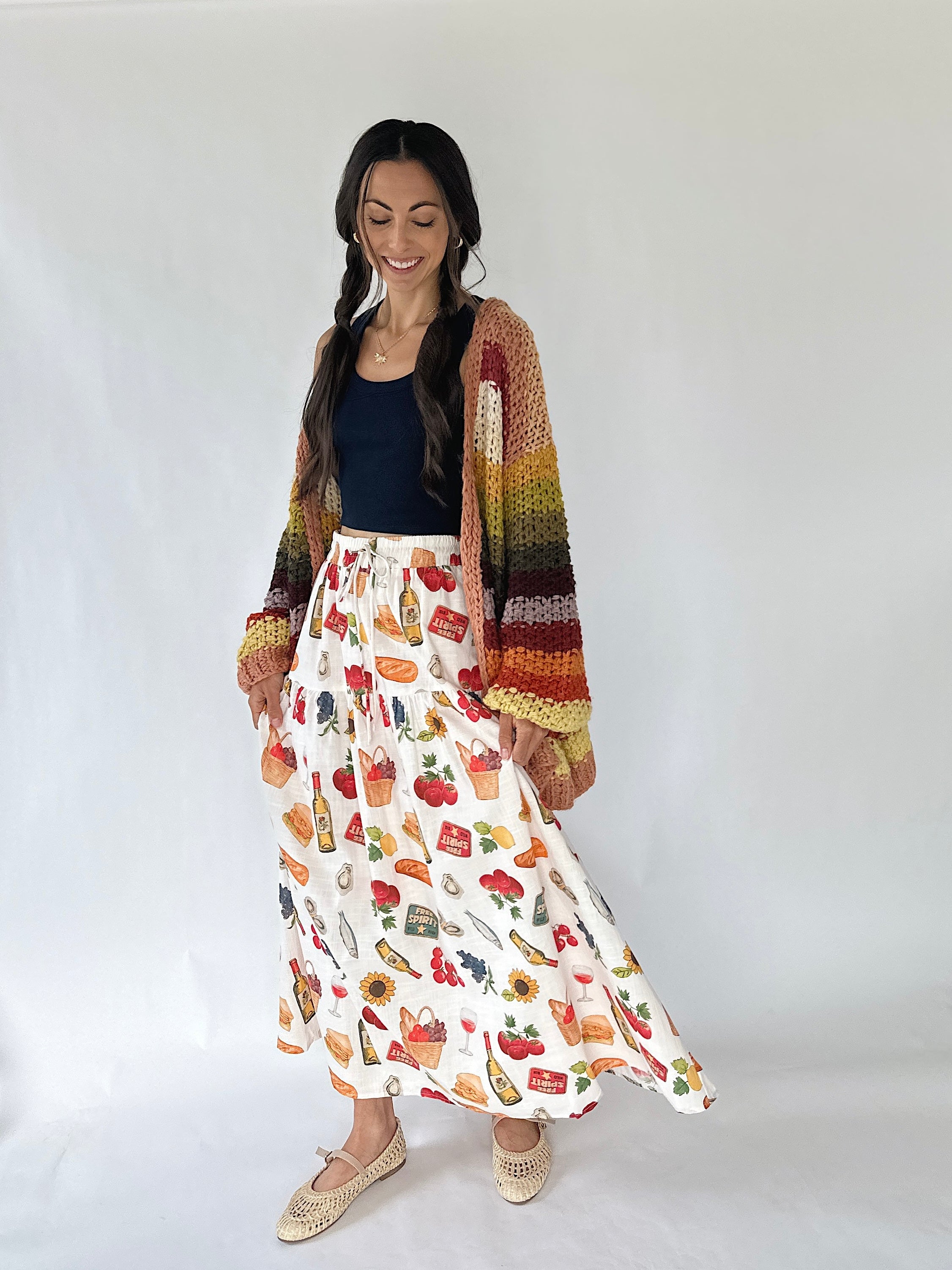 A woman modeling a bohemian-style outfit consisting of a cream maxi skirt with a colorful food print, a dark navy blue tank top, and a multicolor striped crochet cardigan, set against a plain white studio background.