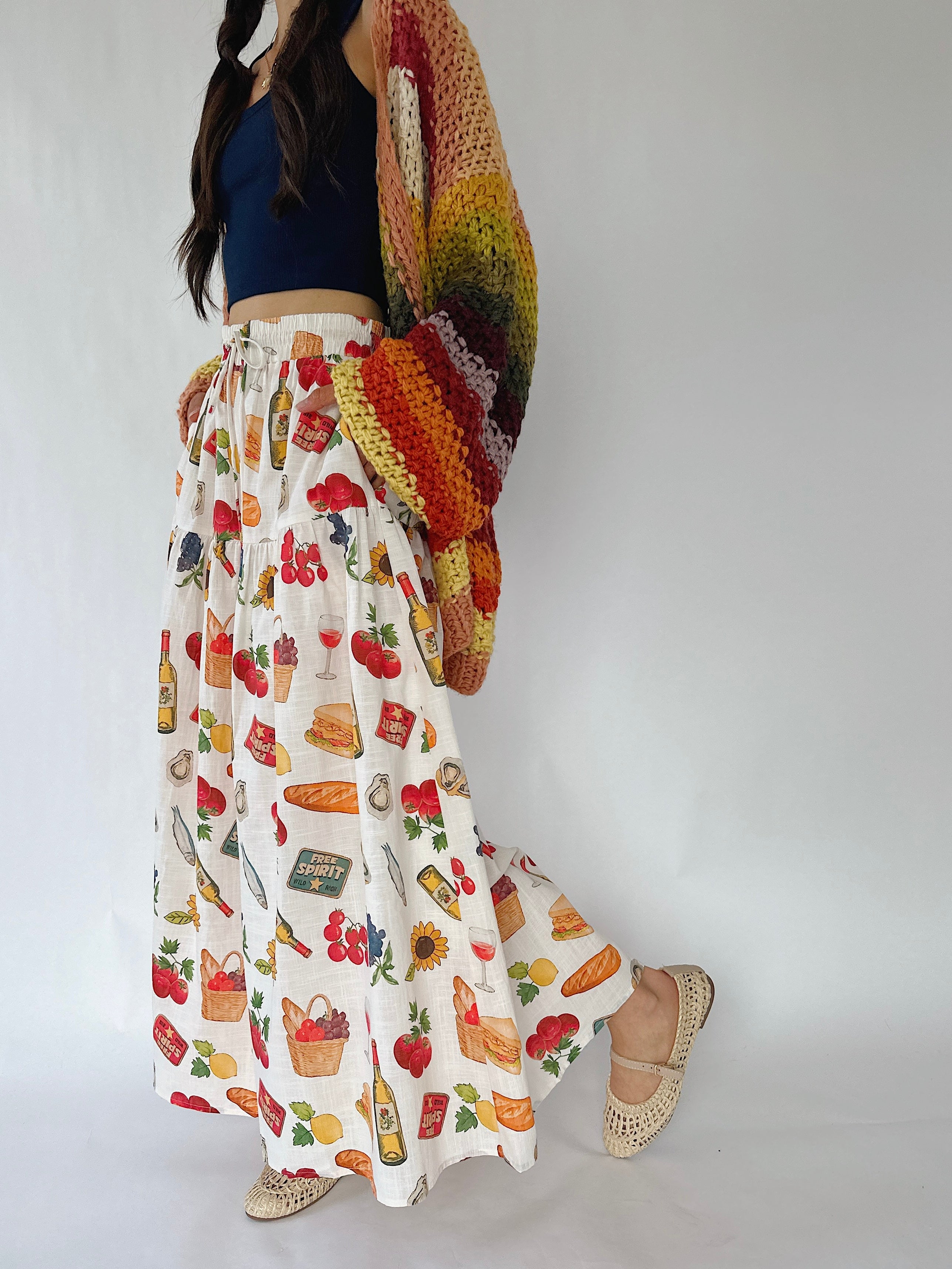 A woman wearing a long white maxi skirt with a colorful fruit and vegetable print, a navy blue crop top, and a chunky multicolor crochet shawl draped over her shoulder, paired with beige woven flat shoes, set against a plain white studio background.