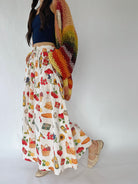 A woman wearing a long white maxi skirt with a colorful fruit and vegetable print, a navy blue crop top, and a chunky multicolor crochet shawl draped over her shoulder, paired with beige woven flat shoes, set against a plain white studio background.