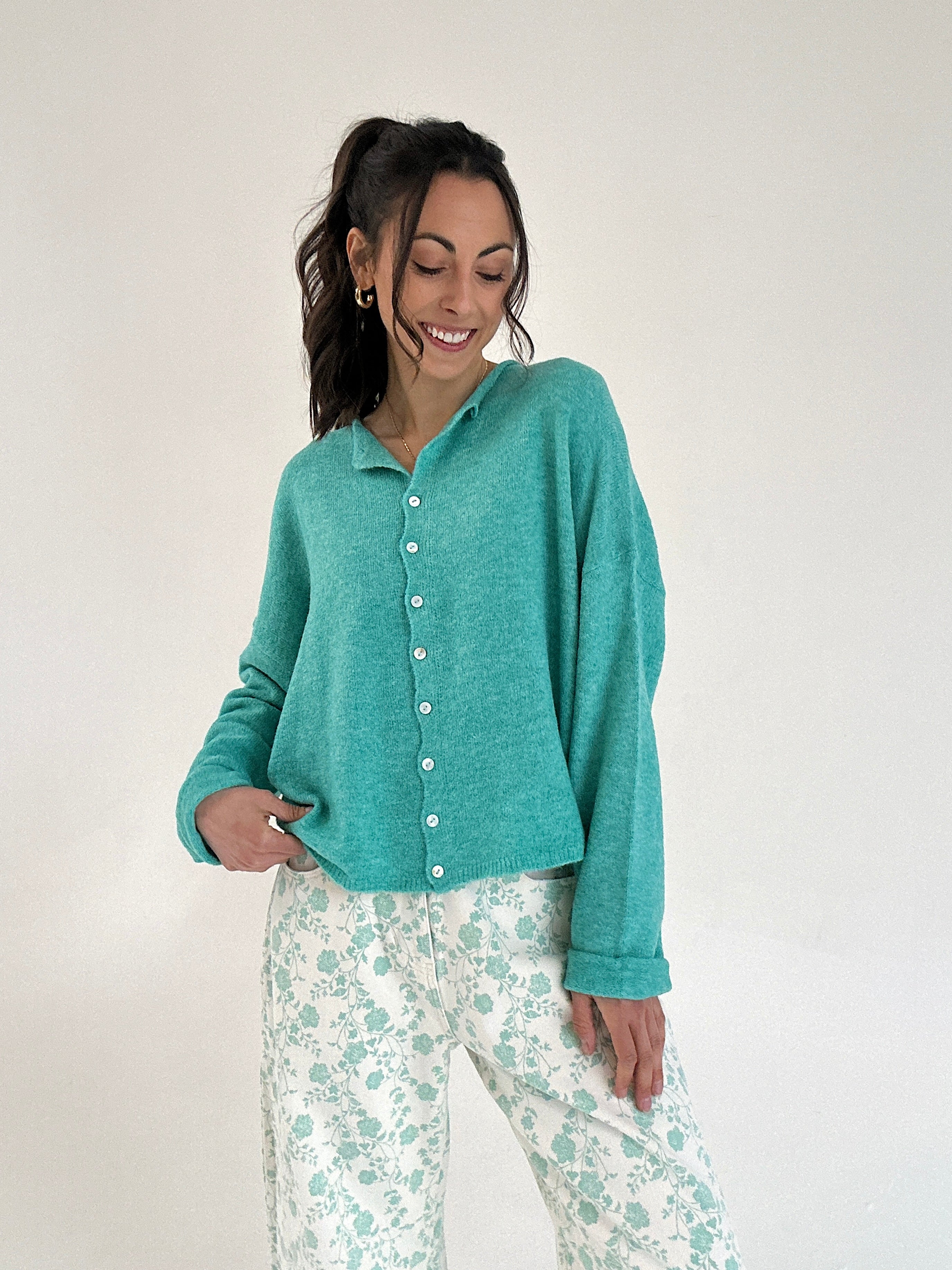 Woman wearing a teal cardigan over white pants with green floral patterns on a plain background