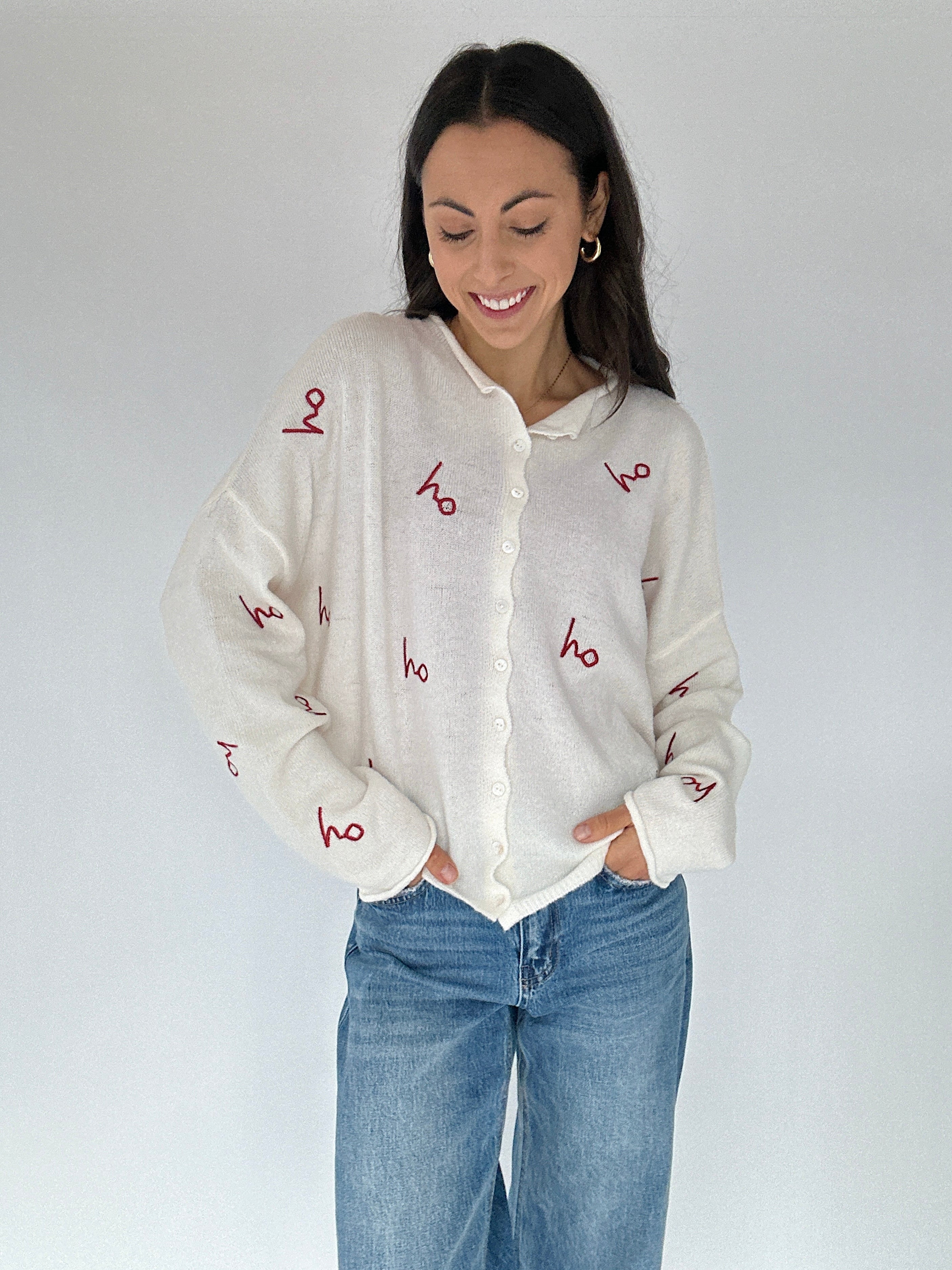 Woman wearing a white cardigan sweater with red embroidered 'ho ho ho' text and blue jeans against a plain background