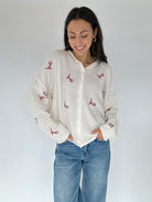 Woman wearing a white cardigan sweater with red embroidered 'ho ho ho' text and blue jeans against a plain background