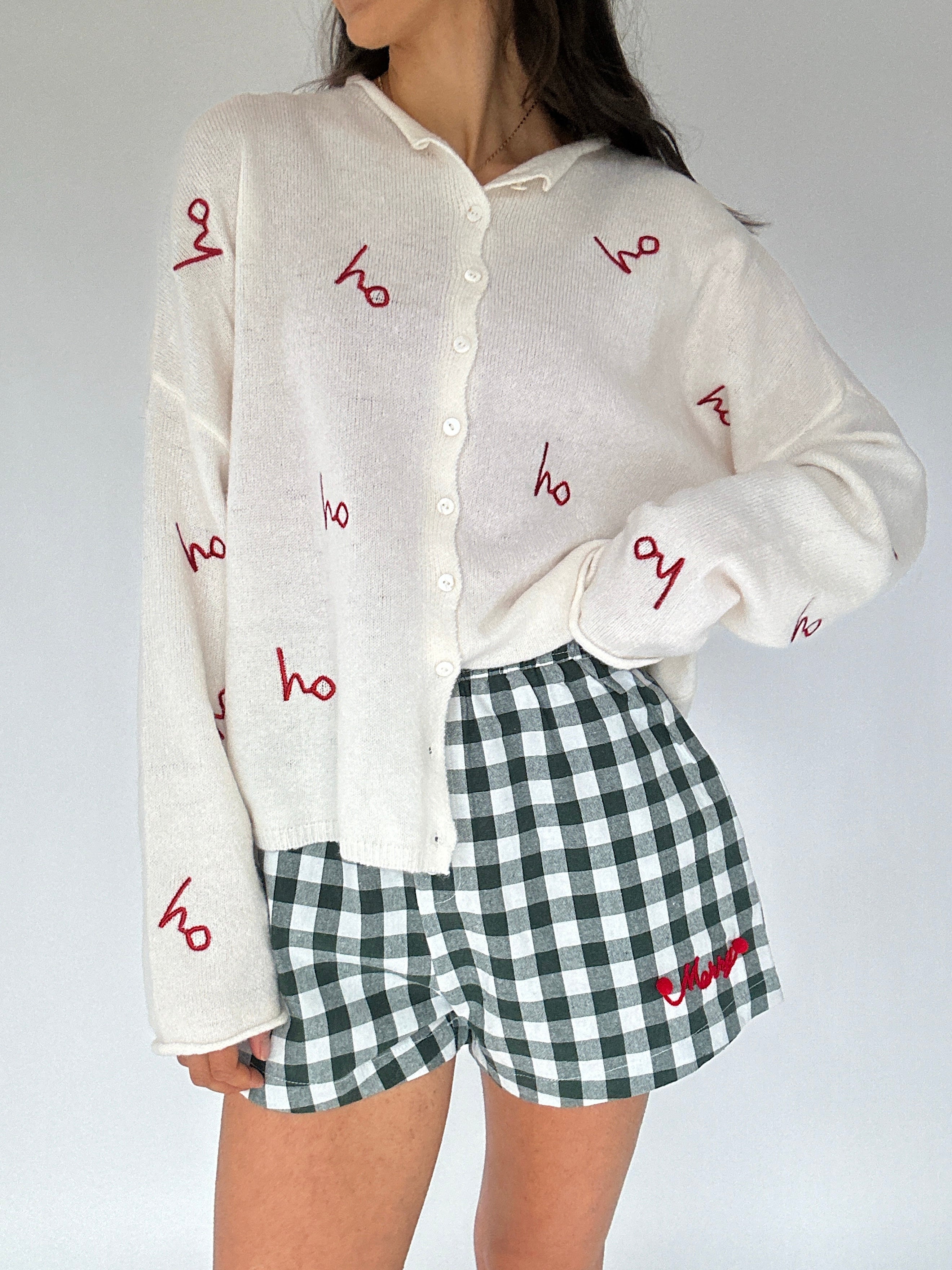 Person wearing a white cardigan with red embroidered 'ho ho ho' text and green and white checkered shorts on a plain background