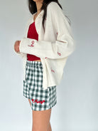 Person wearing a white cardigan with red text, red top, and green checkered shorts on a plain background