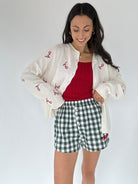 Woman wearing a white cardigan with red text, red sweater tank, and green checkered shorts on a white background