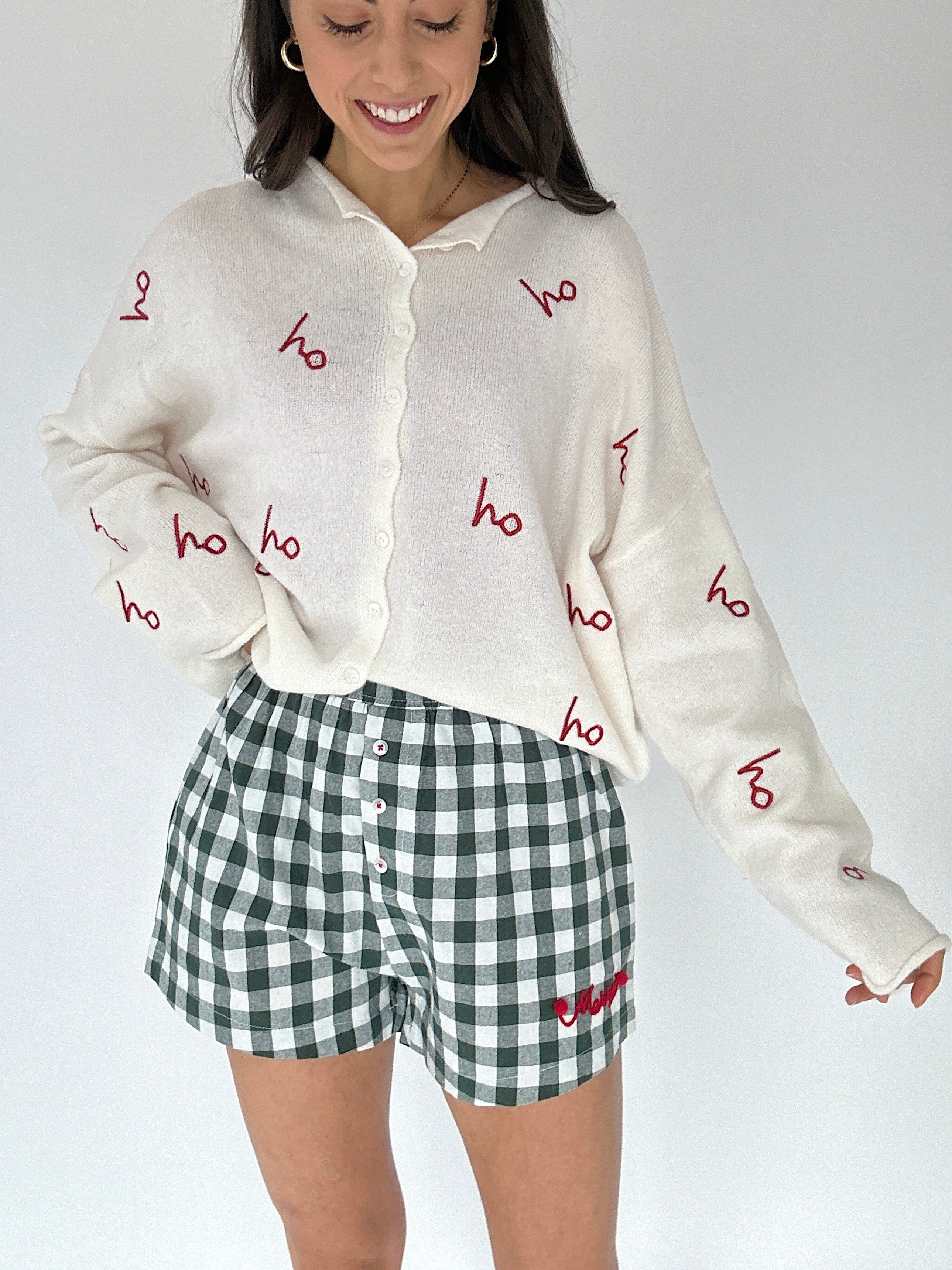 Person wearing a cream-colored cardigan with red 'ho ho ho' text and green checkered shorts on a white background