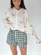 Person wearing a cream-colored cardigan with red 'ho ho ho' text and green checkered shorts on a white background