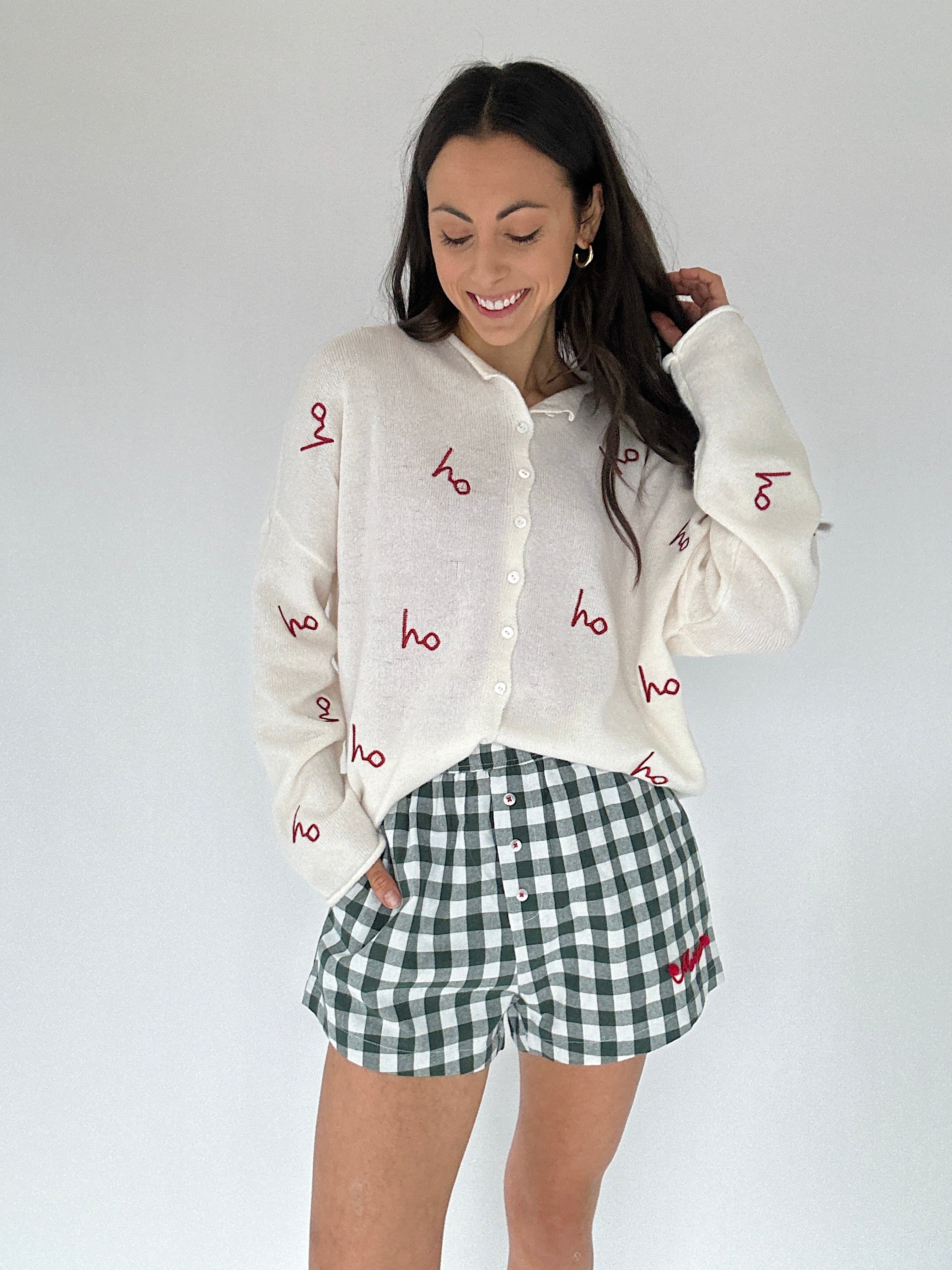 Woman wearing a white cardigan with red text and green checkered shorts on a plain background
