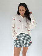 Woman wearing a white cardigan with red text and green checkered shorts on a plain background